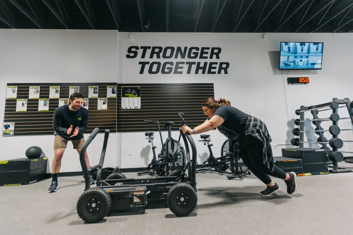 A coach cheers on a member pushing a weighted training sled at an Alloy Personal Training studio